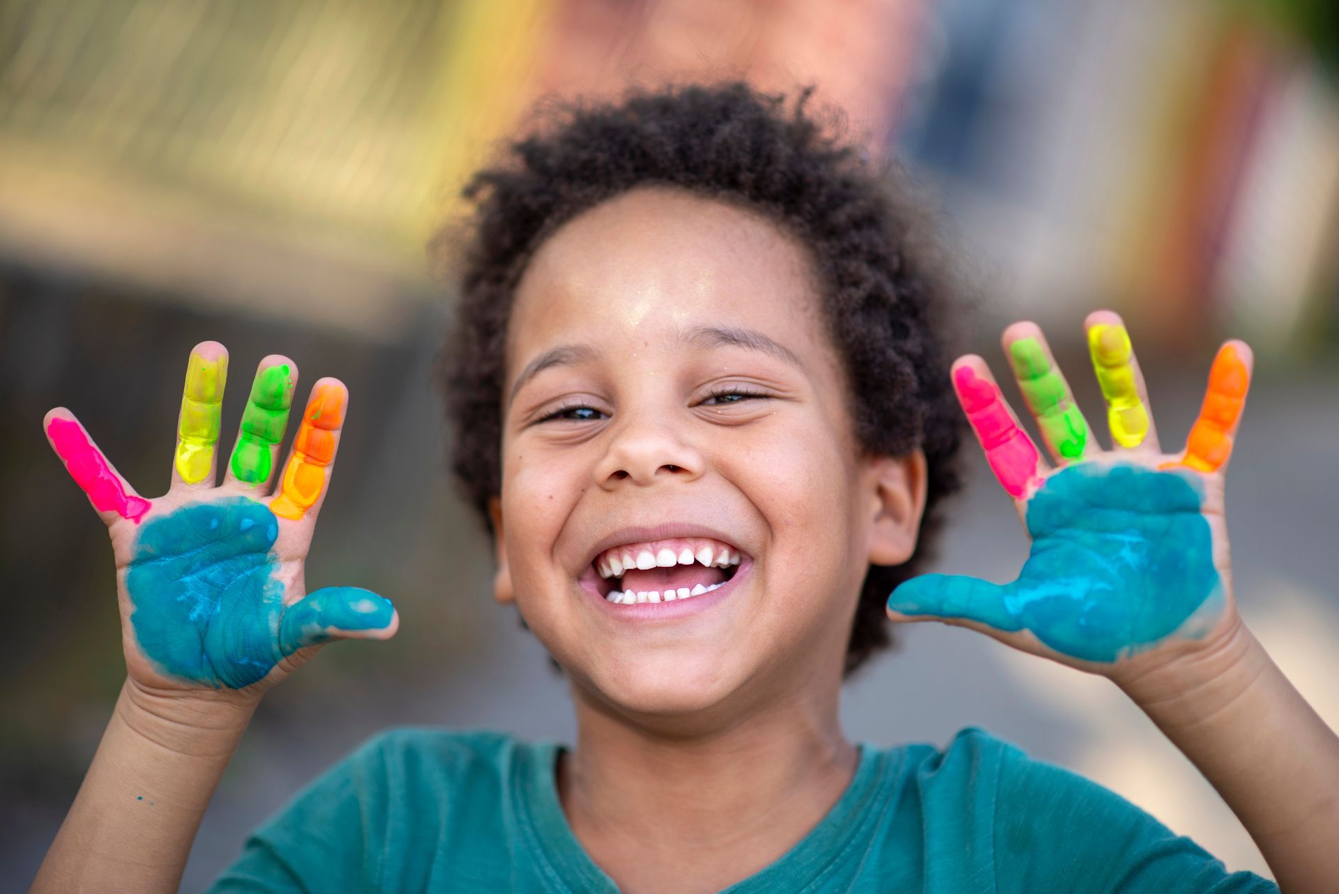 Smiling child showing painted hands with colorful fingerprints against a blurred outdoor background