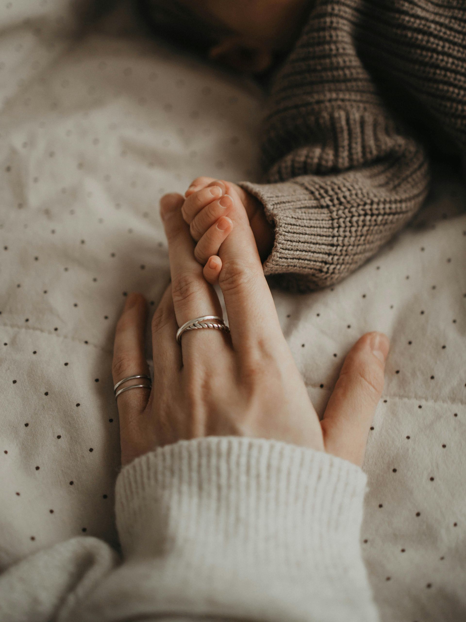 Two hands holding gently on a soft dotted blanket, wearing cozy knit sweaters and rings