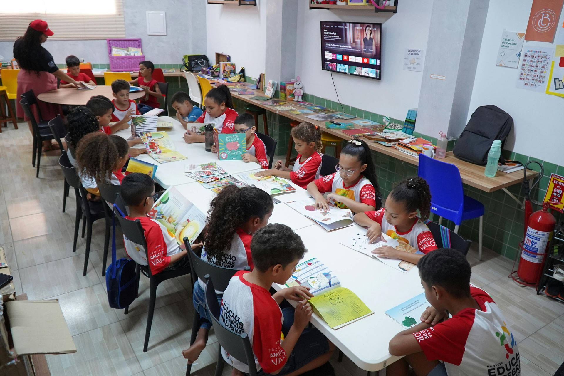 Students in red uniforms sit at tables doing classroom activities while a teacher assists nearby.