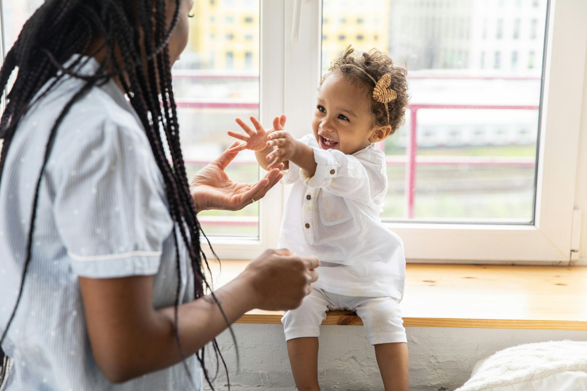 Adult and toddler playing by a bright window, reaching hands toward each other.