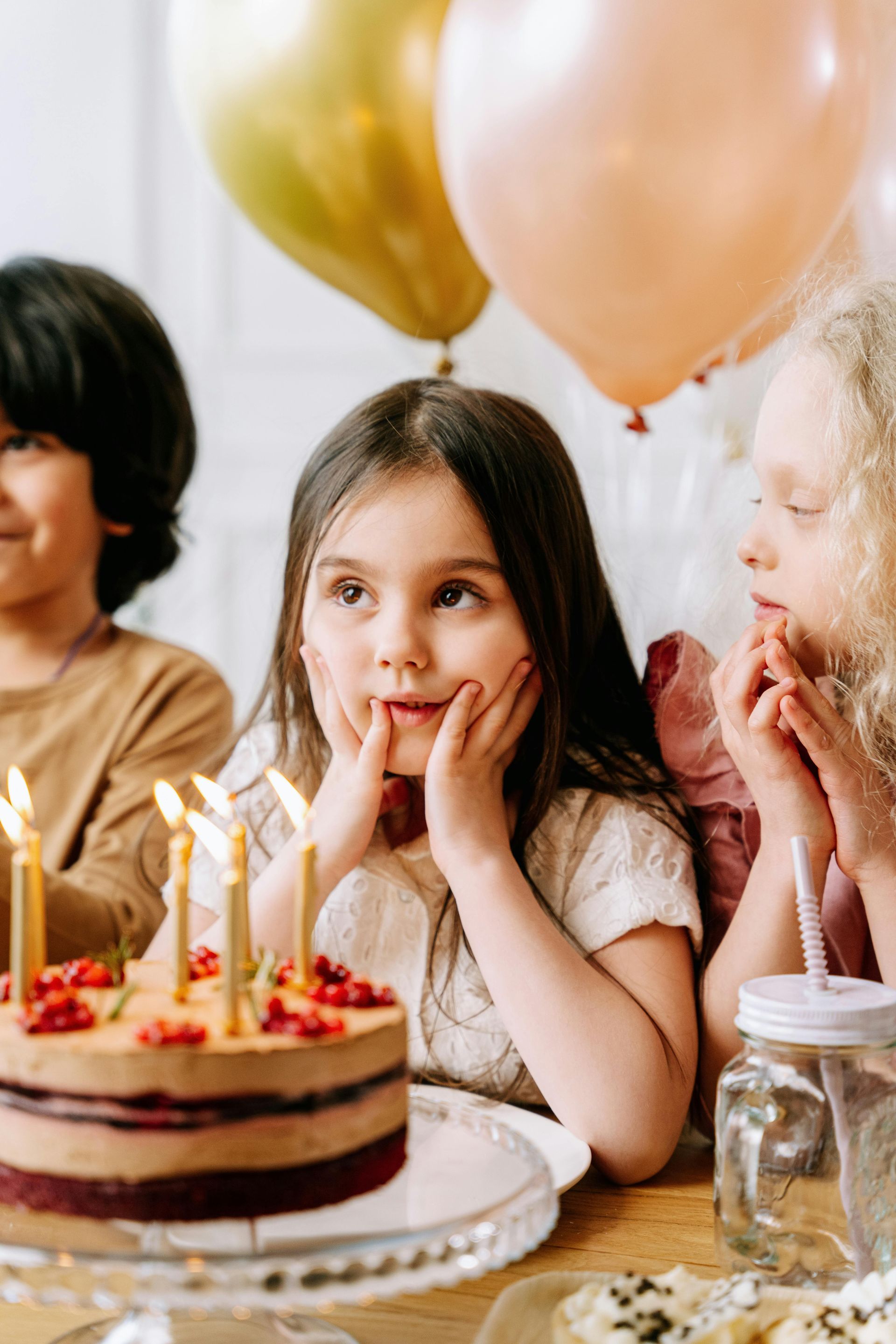 Children at a birthday table, gazing at a cake with lit candles and balloons overhead.