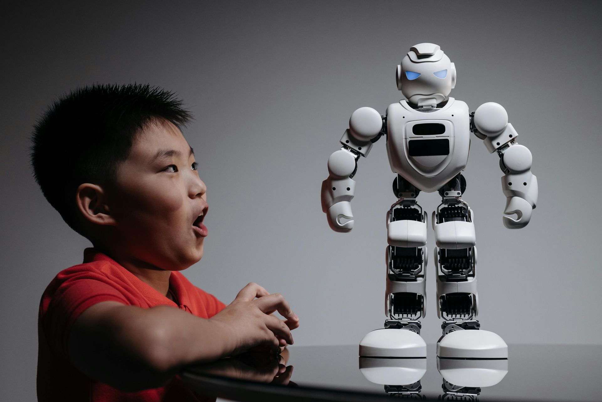 Boy in red shirt looking surprised at a white robot toy on a black table.