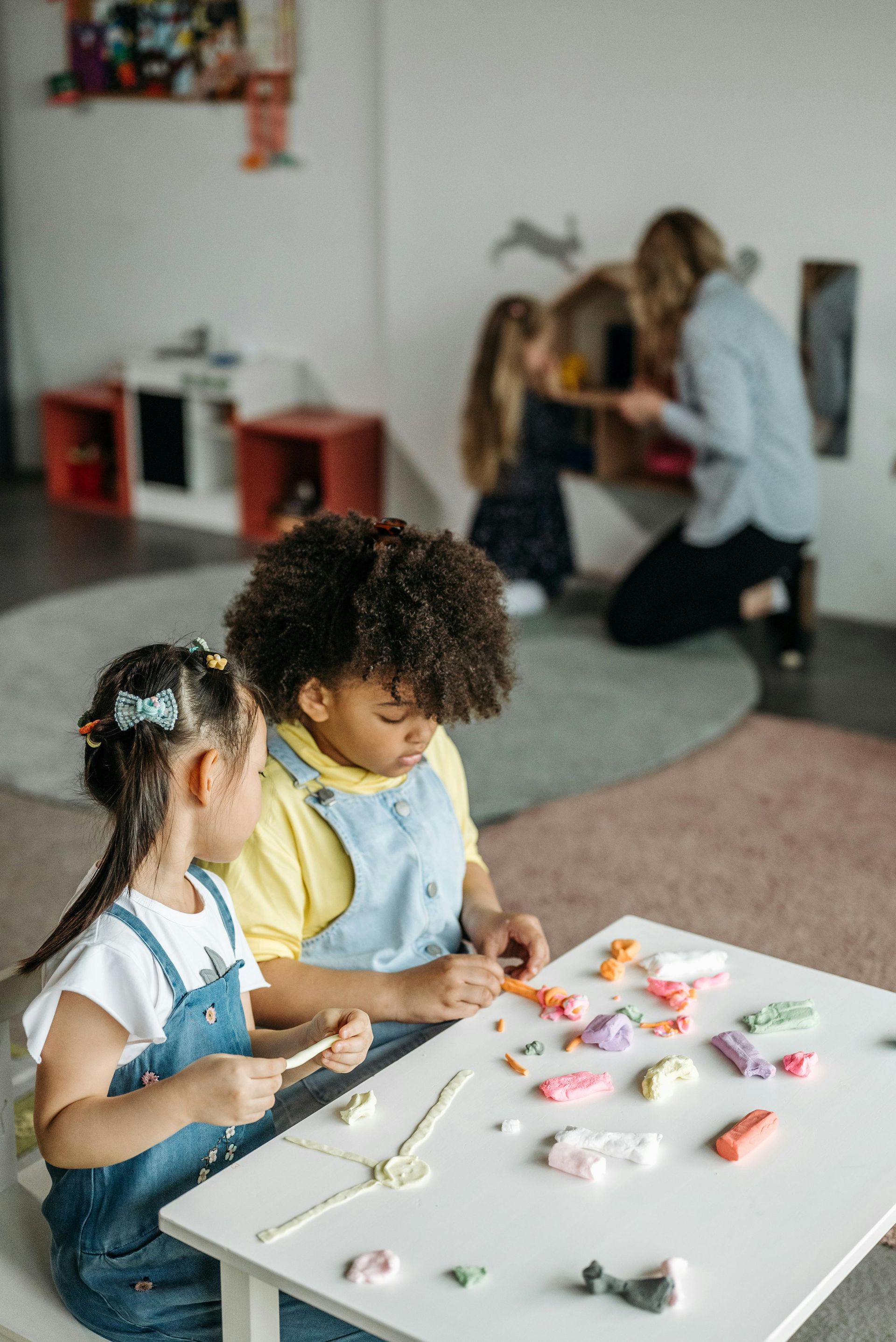 Two children crafting at a white table while an adult helps in the background.