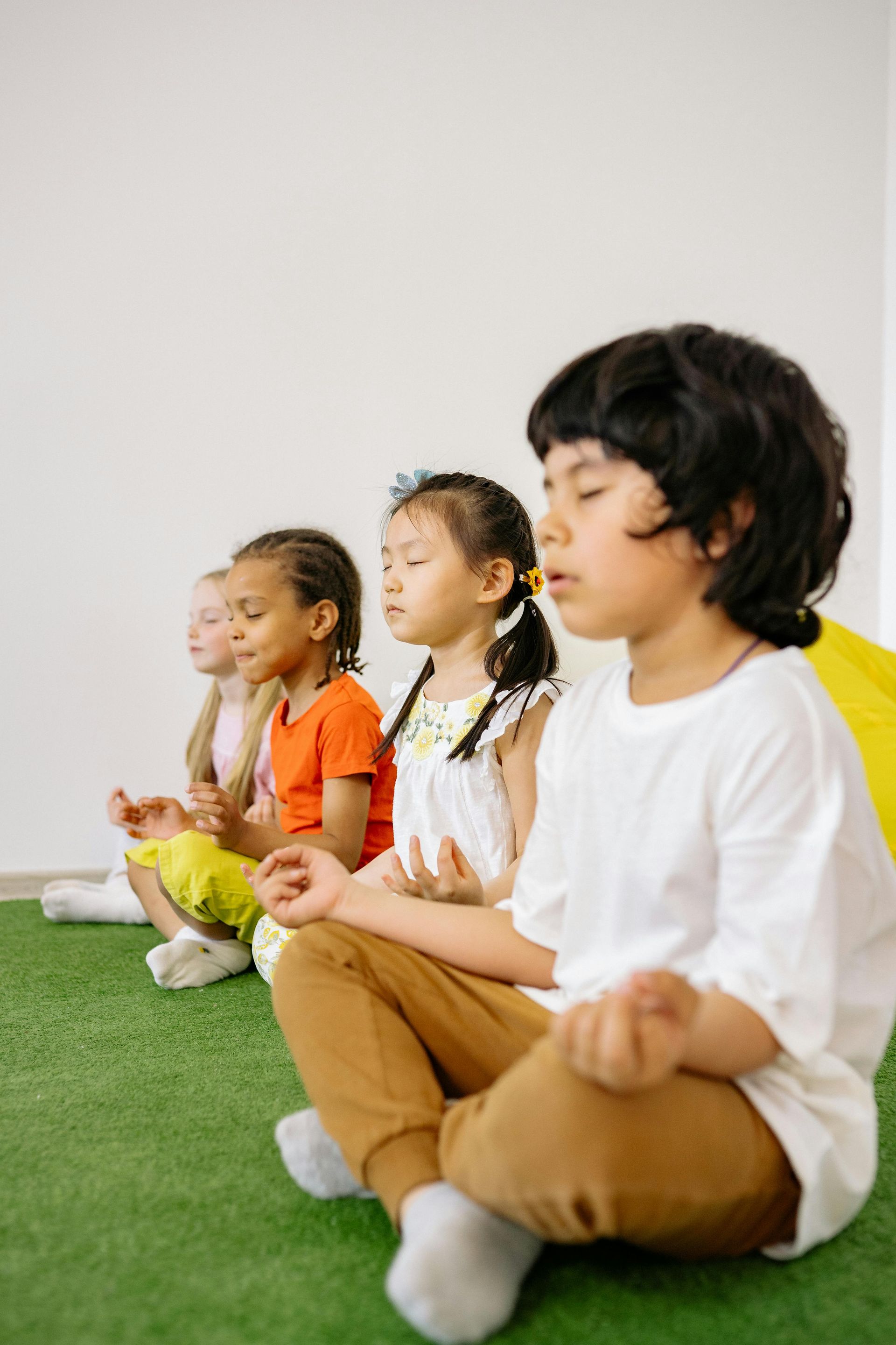Three children sit cross-legged on a green floor against a white wall, meditating in a row.