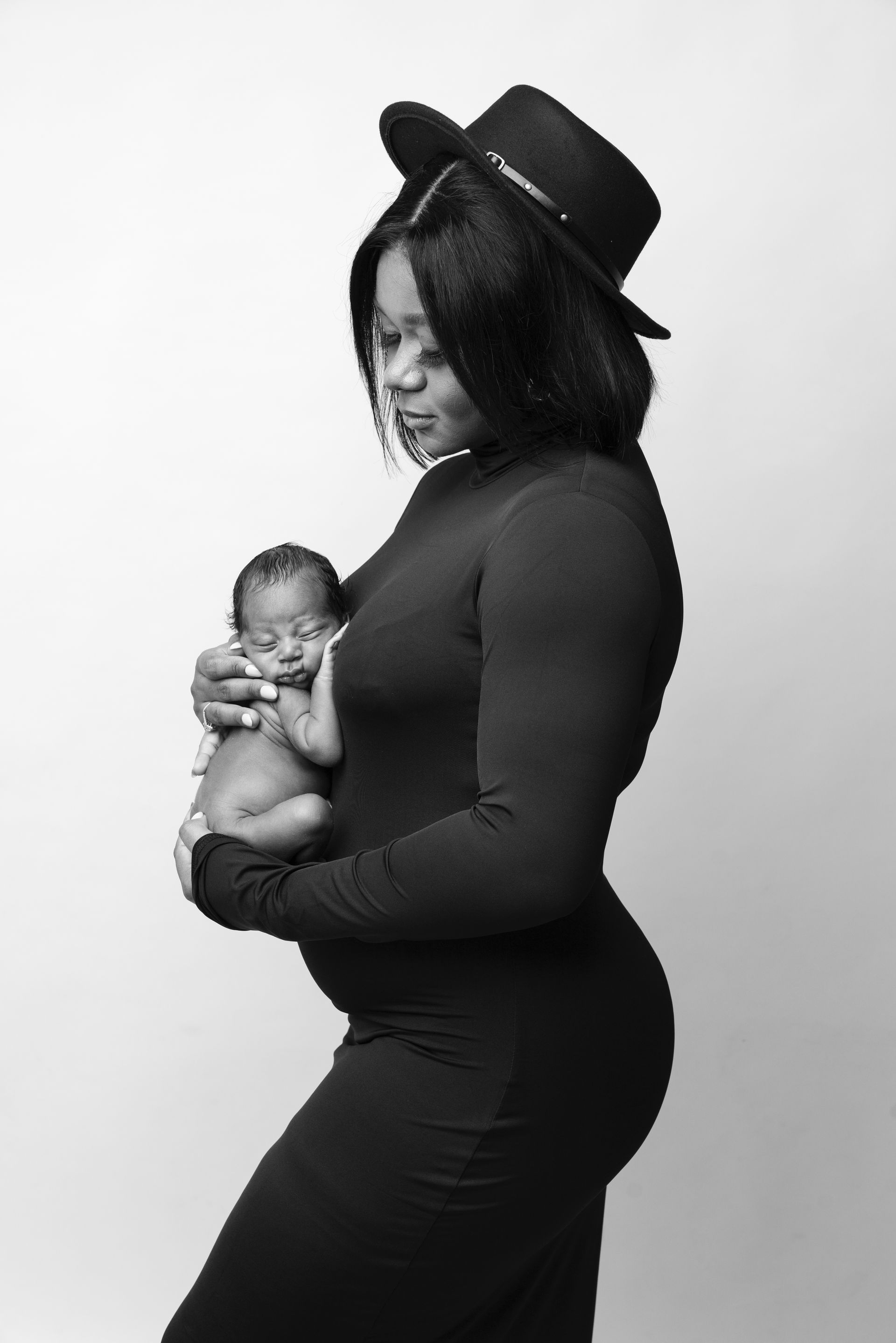 Pregnant person in a black dress and hat gently holding a baby against a plain light background