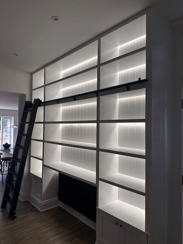 White built-in shelving with LED strip lighting, cupboards below and a black rolling library ladder