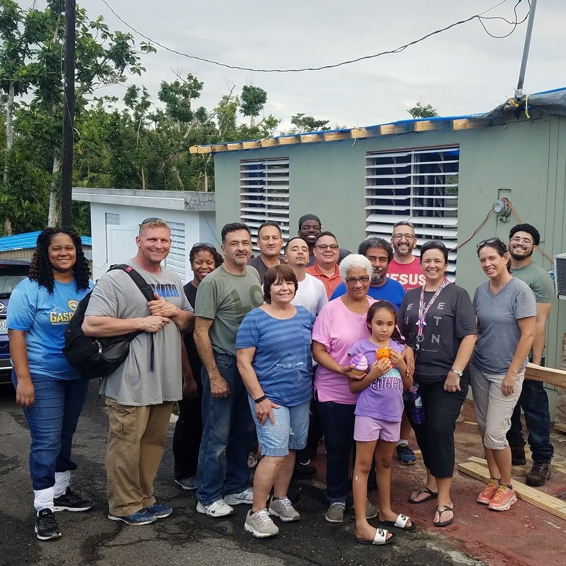 Group of people posing outdoors in front of a small building, smiling and standing close together.