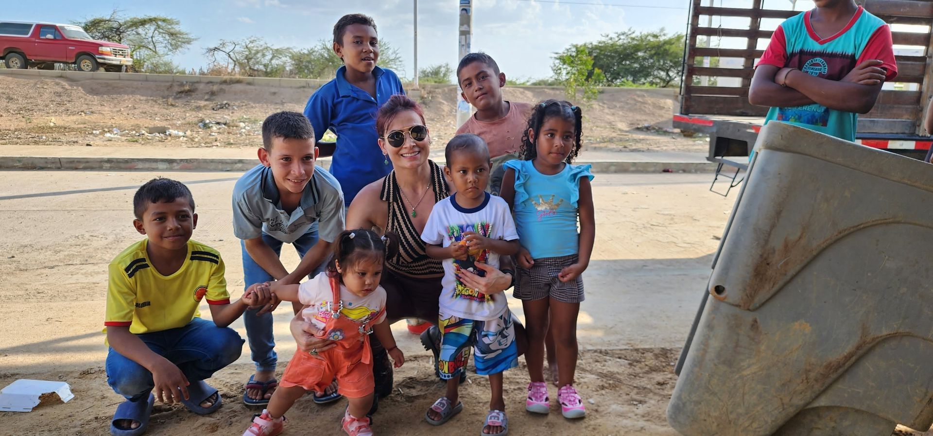 Group of children and two adults posing outdoors beside a large metal container in a sunny rural area