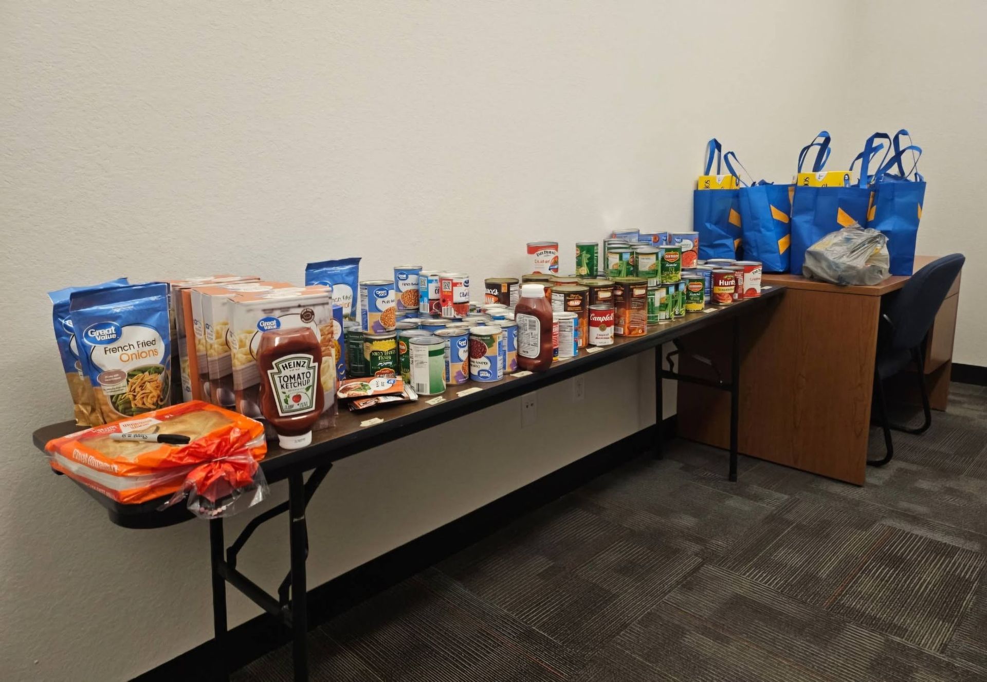 Long table of assorted packaged snacks and drinks beside blue tote bags in a room