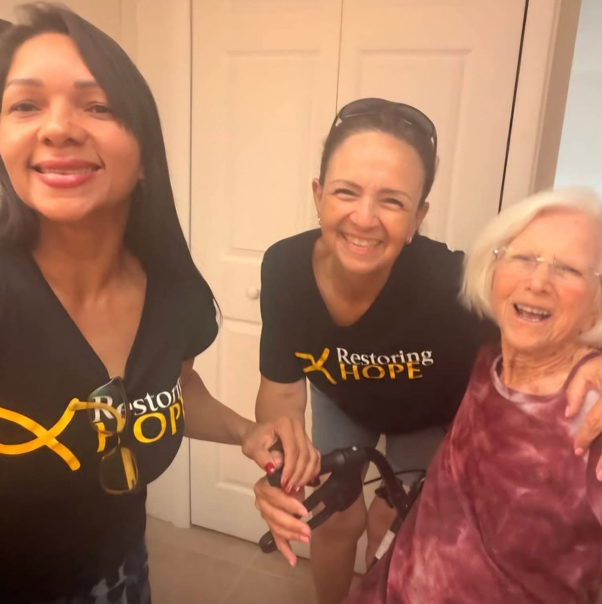 Three women smiling indoors, two wearing Restore Hope shirts