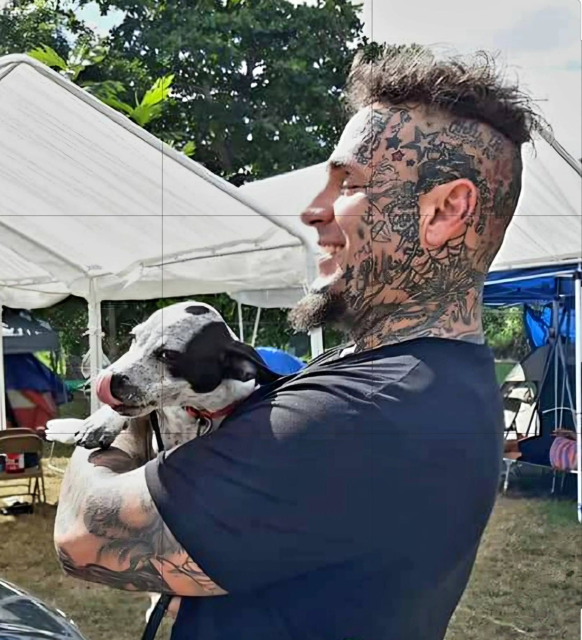 Tattooed man holding a black-and-white dog outdoors under white tents