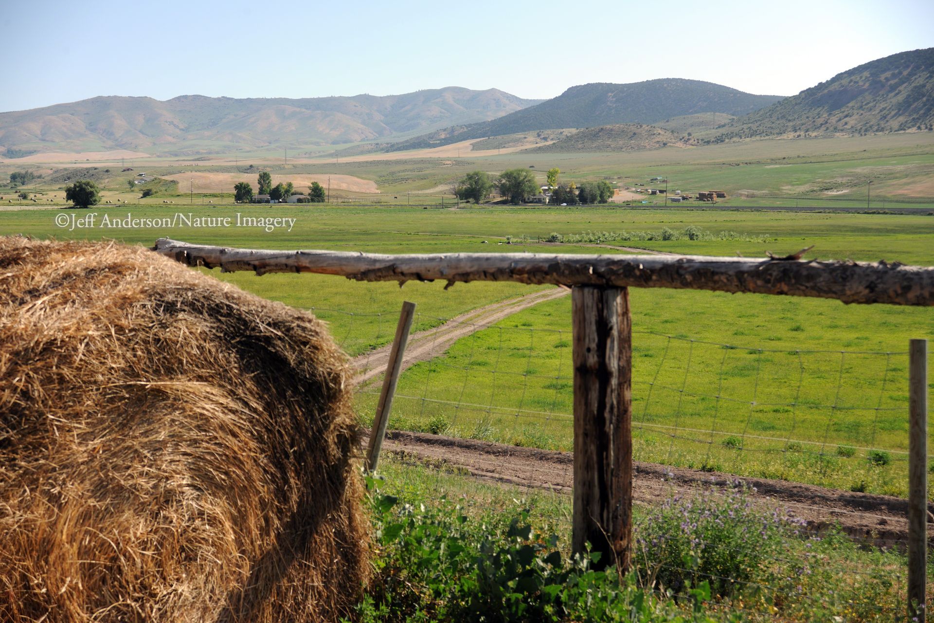 A hay bale sits in the foreground beside a rustic wooden fence overlooking a vast, green valley and rolling mountains.