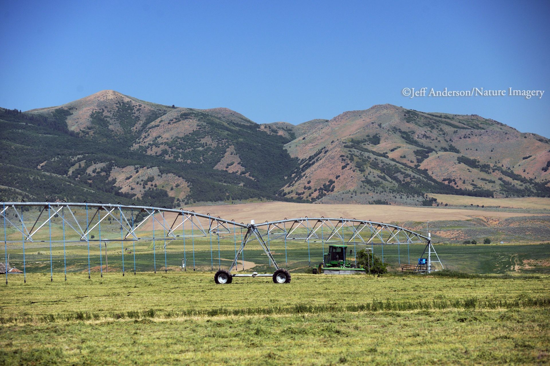 Center pivot irrigation equipment in a green field with mountains in the background under a clear blue sky.