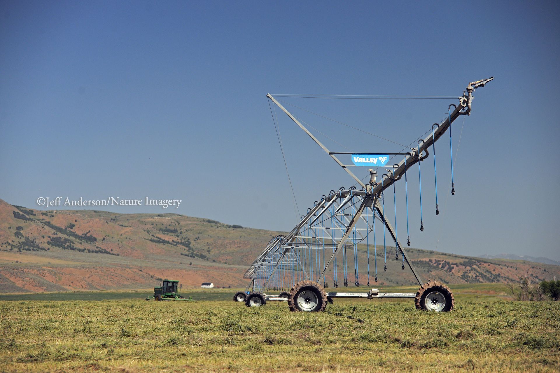 A large metal irrigation pivot stands in a harvested field with rolling hills and a clear blue sky in the background.