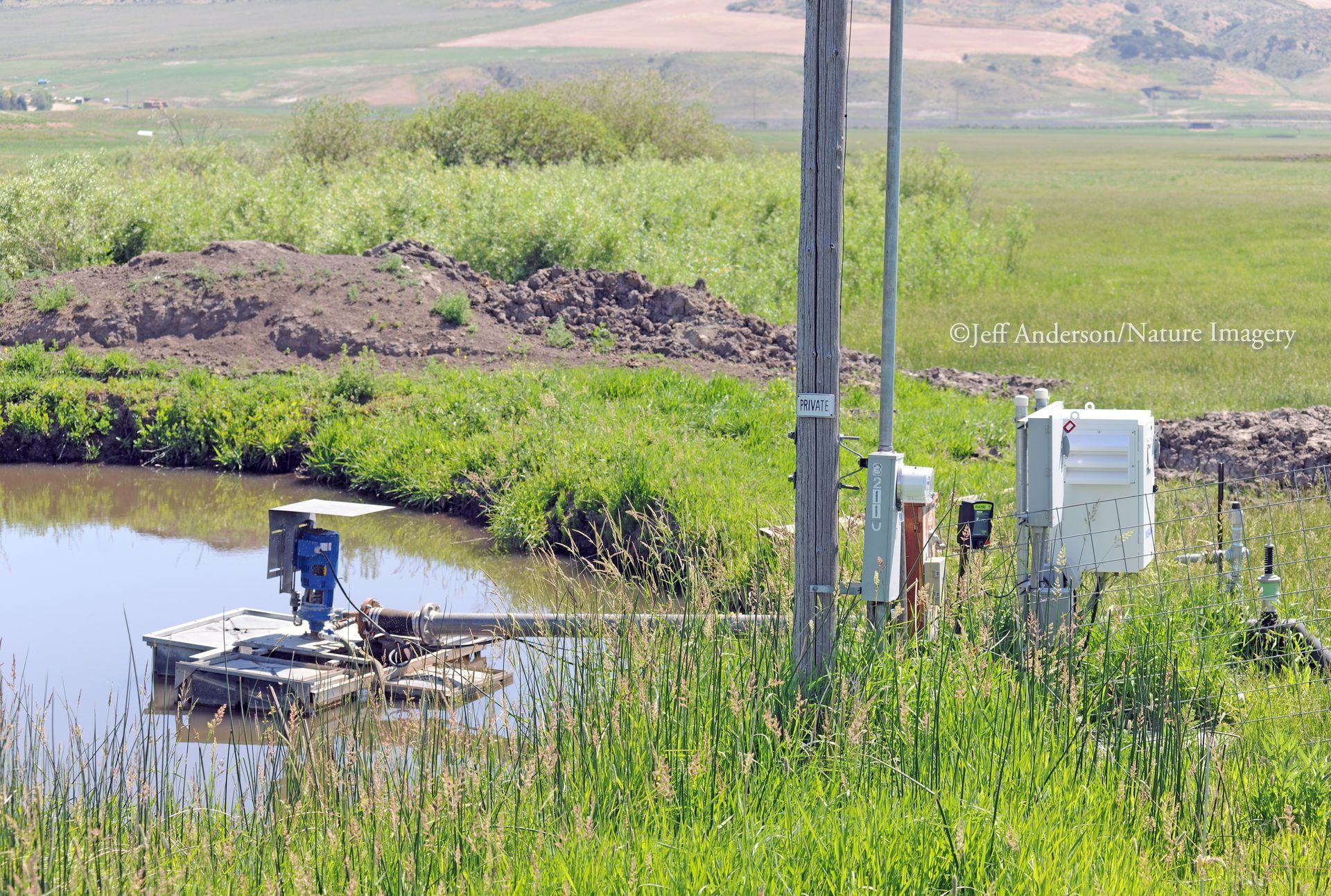 A floating pump mechanism sits in a pond next to electrical control boxes and a utility pole in a grassy landscape.