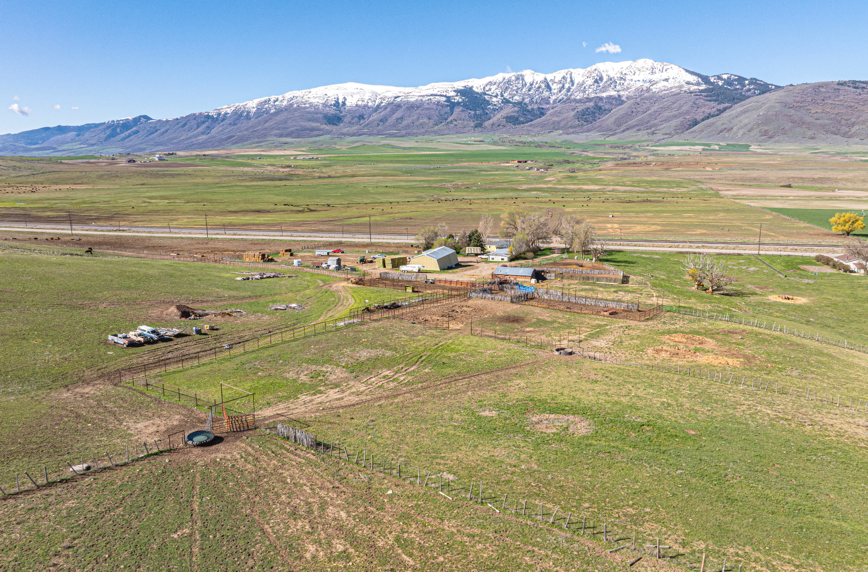 An aerial view of a small rural property with farm buildings set in a vast green valley before snow-capped mountains.