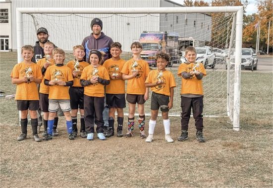 Youth soccer team posing in orange jerseys in front of a goal on a grassy field, with coaches behind them.