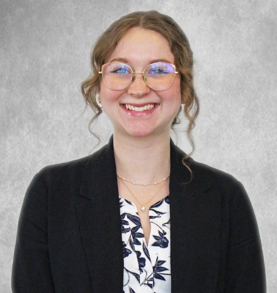 Smiling woman in glasses wearing a black cardigan over a patterned blouse against a gray backdrop