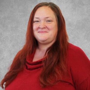 Portrait of a person with long red hair wearing a red top against a gray background