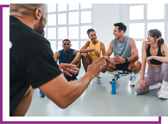 Group exercising and talking in a bright studio, with a trainer in the foreground.
