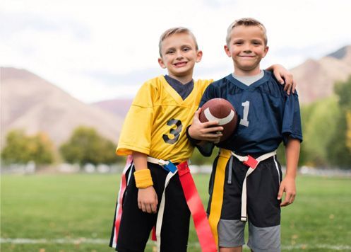 Two children in football gear standing on a field, one holding a football and both smiling.