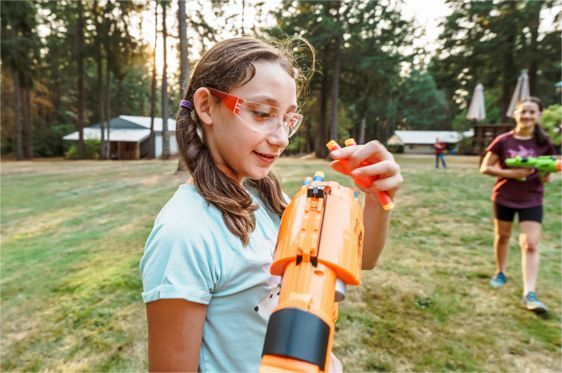 Girl in clear goggles inspects an orange water rocket launcher at a grassy outdoor field.