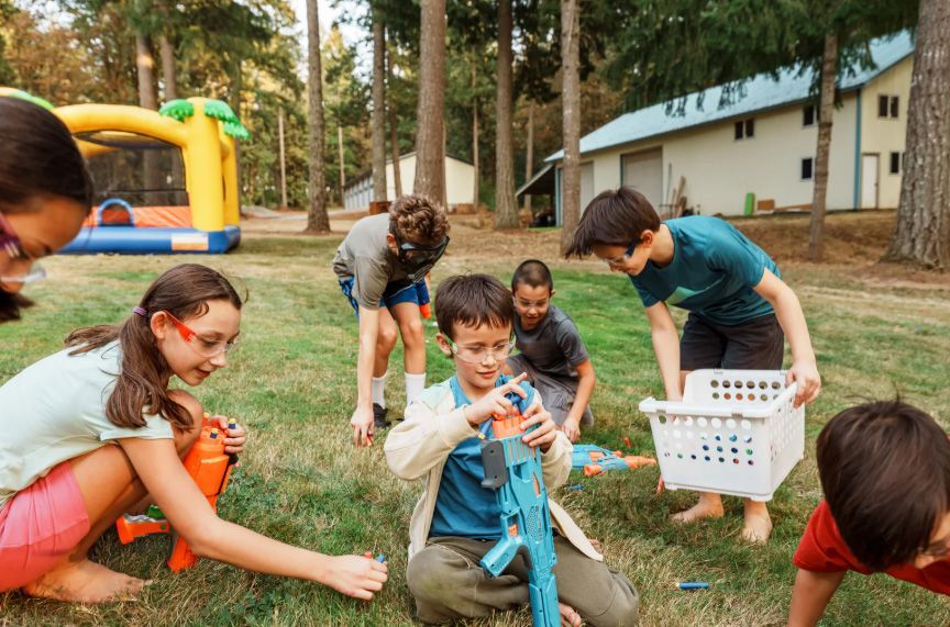 Children playing with bubbles on a grassy lawn outside a building, with a bucket and inflatable playground in the background