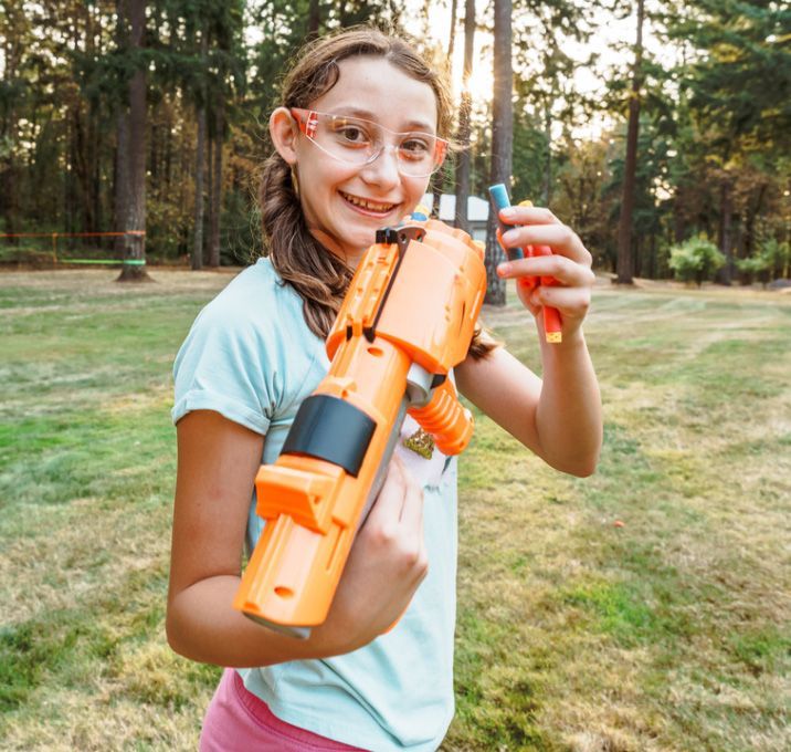 Child holding an orange toy blaster outdoors, smiling in a grassy yard.