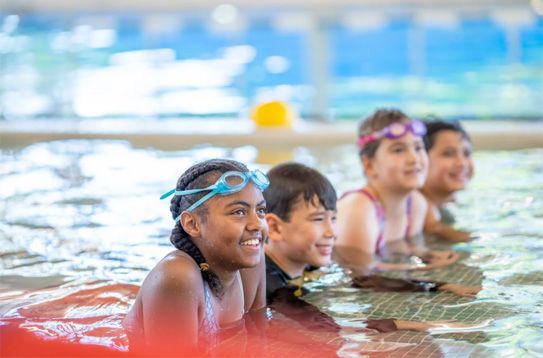 Children in a pool, smiling and holding the edge with swim goggles on