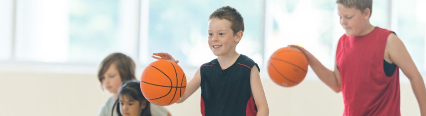 Children playing basketball indoors, smiling while dribbling orange balls