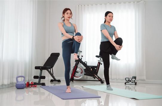 Two people exercising on yoga mats in a bright home studio with a stationary bike and dumbbells nearby