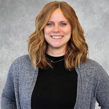 Smiling person with wavy blonde hair in a gray cardigan and black top against a gray background