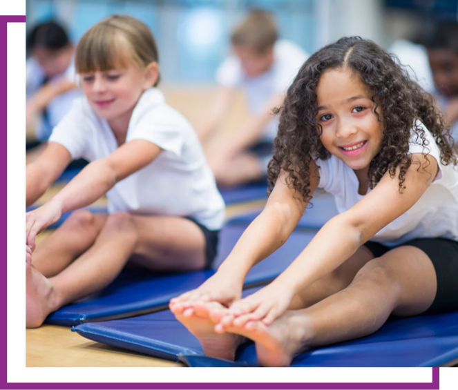 Children stretching on blue mats in a gym class, smiling toward the camera.