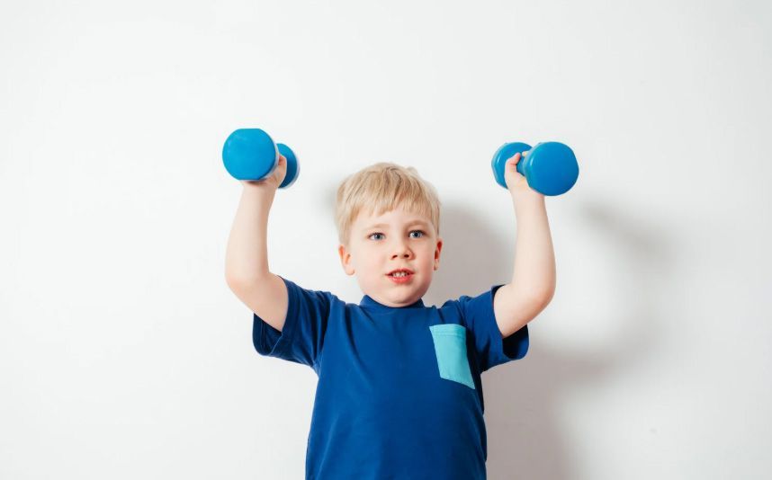 Child lifting blue dumbbells against a white wall, wearing a blue shirt.