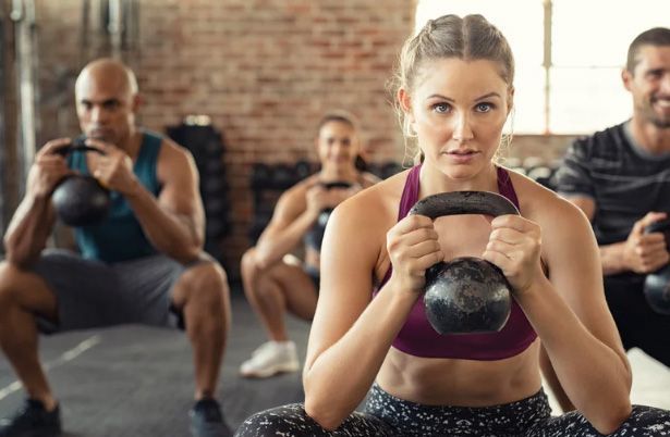Group exercise class doing kettlebell squats in a gym, with a woman centered in front holding a black kettlebell.