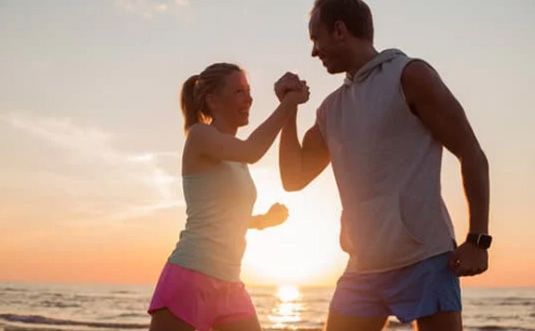 Two people high-five on a beach at sunset, with the ocean and warm sky behind them