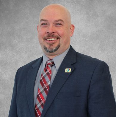 Smiling bald man in a navy suit, striped tie, and lapel pin against a gray background