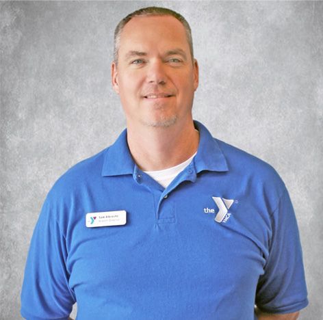 Man in a blue YMCA polo shirt with a name tag, standing against a gray backdrop.
