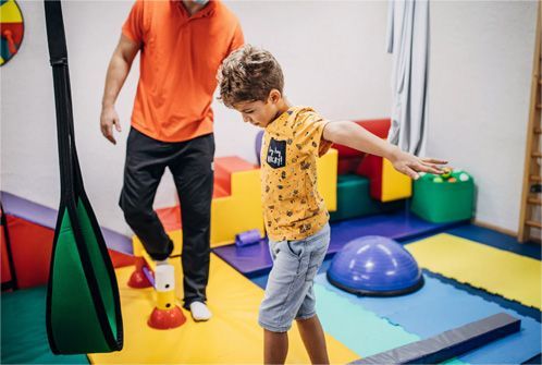 Child balancing on a blue mat in a colorful therapy room with an adult nearby