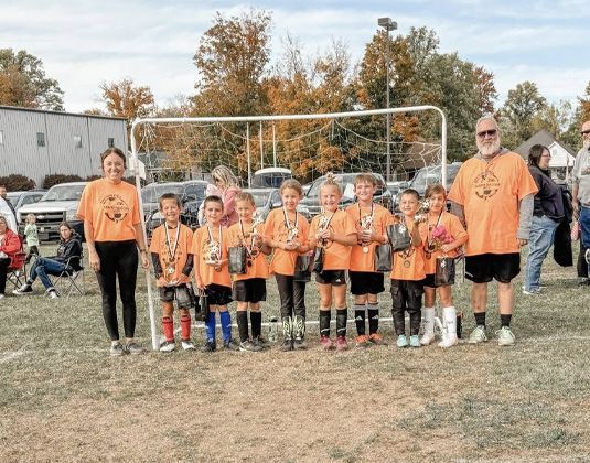 Youth soccer team in orange jerseys posing together on a field in front of the goal, with coaches on both sides