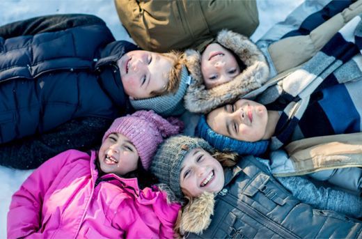 Five children in winter coats and hats lie close together on blankets, smiling up at the camera.