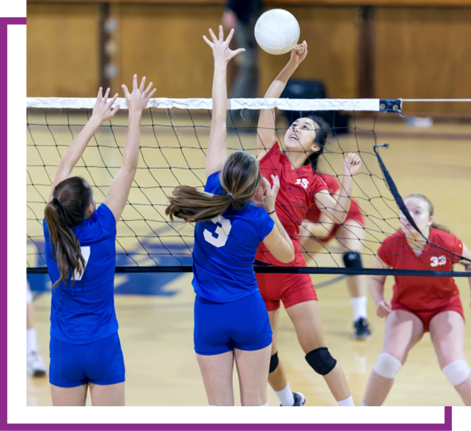 Volleyball players in red and blue jump at the net for an overhead hit during a gym match