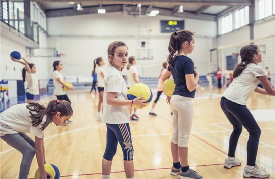 Children practicing with dodgeballs in a gymnasium, holding yellow and orange balls.