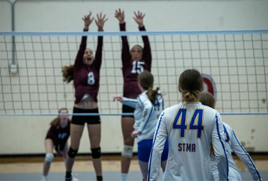 Volleyball players at the net; two maroon blockers jump as a player in a white jersey watches from the foreground