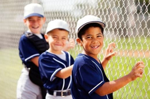 Three children in blue uniforms and white caps smiling by a chain-link fence outdoors