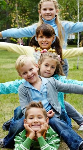 Four children forming a line in a park, each smiling with arms outstretched, standing in a lush green outdoor setting.