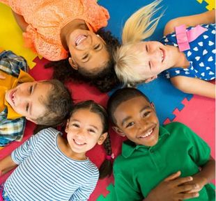 Five children lying in a circle on a colorful foam mat, smiling and looking up toward the camera.