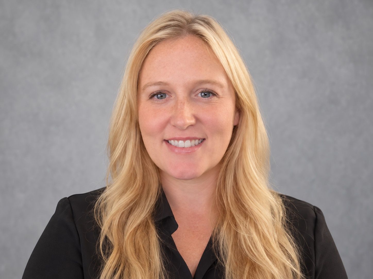 Smiling blonde woman in a black blazer against a gray backdrop
