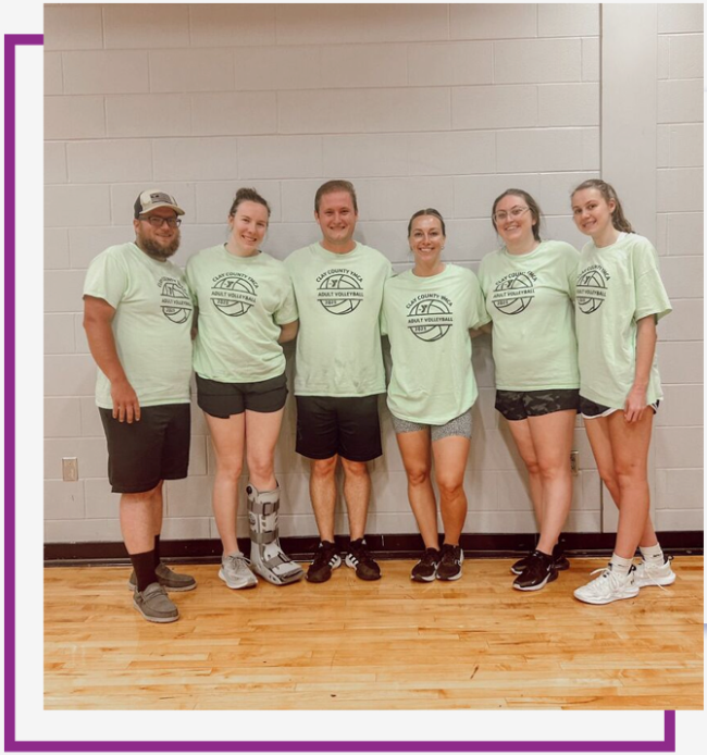 Six people in matching light green shirts stand together in a gym against a white brick wall.