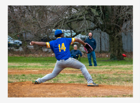 Baseball pitcher in blue jersey and gray pants throws from the mound toward a catcher and umpire behind him